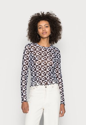 Camisola de mangas longas em tecido transparente, com um padrão de coração e tabuleiro de xadrez em branco e castanho, apresentando uma bainha assimétrica. Combinada com jeans brancos.
