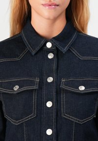 Kvinne iført mørk denimjakke med sølvfargede trykknapper og frontlommer, vist fra nakken til midt på torso.