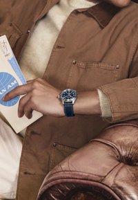 Analogās pulkstenis ar tumši zilu ciparnīcu, sudraba toņa korpusu un zilu ādas siksniņu. Roku tur salocītu ceļojuma biļeti pie brūnas jakas.