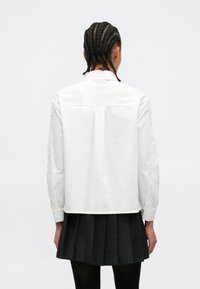 Λευκό πουκάμισο με δαντέλα στα μανίκια, μονόχρωμη πλάτη και μανσέτες με κουμπιά. Συνδυασμένο με πλισέ μαύρη φούστα.