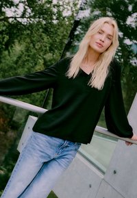 Schwarzes Longsleeve-Oberteil mit subtiler Textur und V-Ausschnitt kombiniert mit hellblauen Jeans, vor einem grünen Außenbereich.
