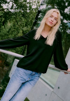 Musta pitkähihaine paita, jossa on hienovarainen rakenne ja V-kaula-aukko, yhdistettynä vaaleansinisiin farkkuihin vihreää ulkoilman taustaa vasten.