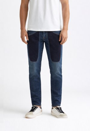 Uomo che indossa jeans blu a due tonalità a taglio slim con toppa scura sulle cosce anteriori, sneakers bianche con lacci neri e una t-shirt bianca semplice.