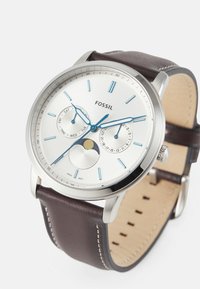 Montre en acier inoxydable argenté avec un cadran blanc, des aiguilles bleues, un indicateur de phase de lune et un bracelet en cuir marron. Comprend un affichage du jour de la semaine.