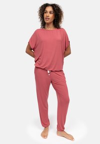 Frau mit lockigem Haar, die lockere rosa Freizeit-Hosen und ein Oberteil trägt, steht barfuß vor einem schlichten weißen Hintergrund.