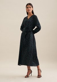 Leopardenmuster Wickelkleid in Dunkelgrün und Schwarz, mit langen Ärmeln, Taillenbindung, fließendem Stoff, knielang, kombiniert mit schwarzen Riemchen-Heels.