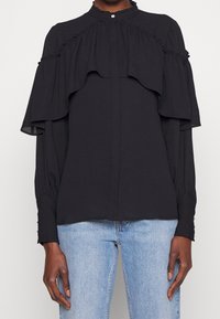 Schwarze Bluse mit geschichteten Rüschendetails, hohem Kragen und Knopfverschluss. Lange Ärmel mit gepufften Manschetten und aus einem glatten, leichten Stoff.