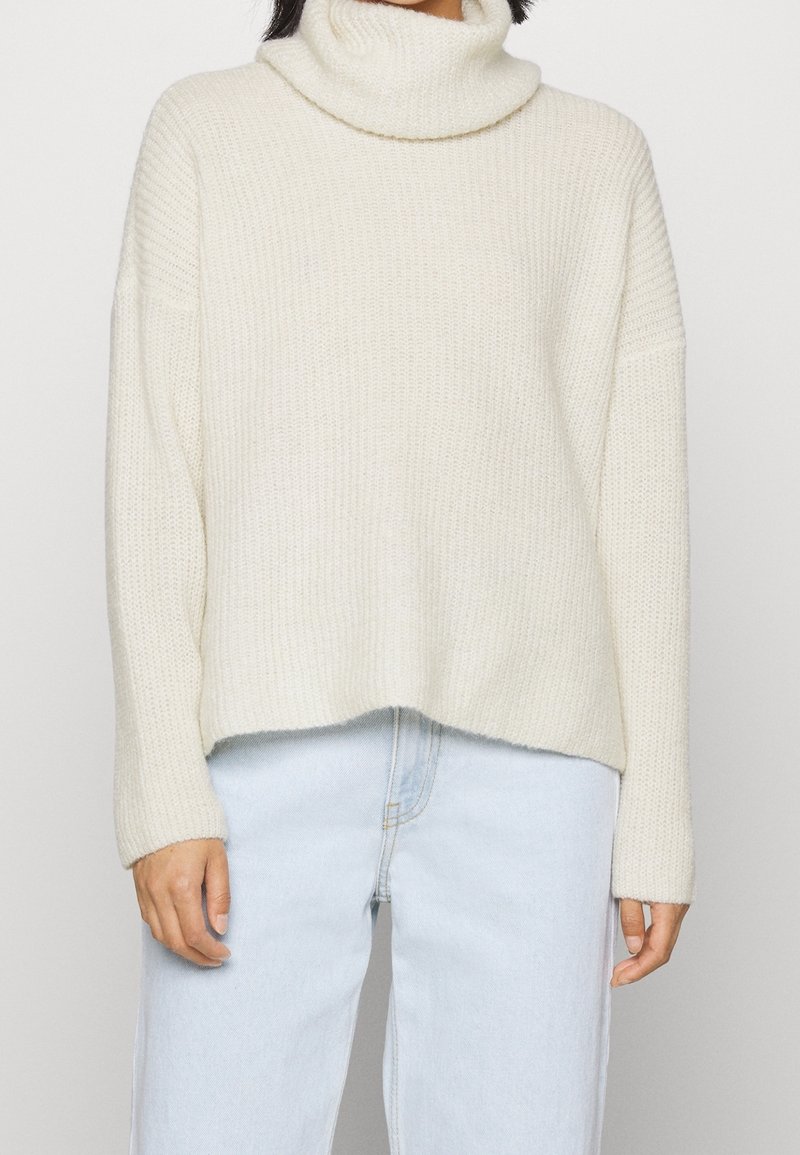 Personne portant un pull à col roulé en tricot crème et un jean bleu clair, debout devant un fond clair uni.