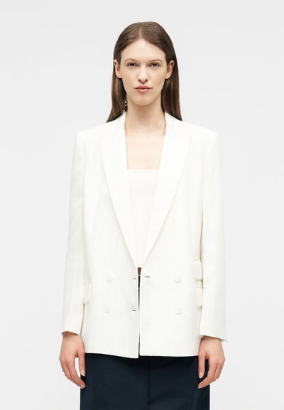 Femme portant un blazer blanc à double boutonnage sur un top blanc et un pantalon foncé, debout devant un fond clair uni.