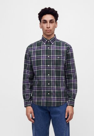 Jeune homme portant une chemise à carreaux verte, violette et blanche boutonnée et un jean bleu, debout devant un fond blanc uni.
