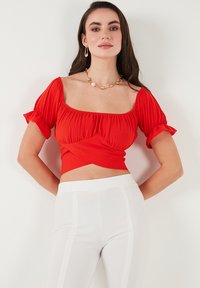 Rød kort top med korte puffærmer, tekstureret stof og design med snoet front, parret med hvide højtaljede bukser med sidelister.