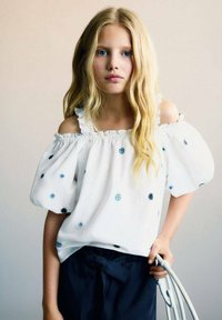 Blusa bianca off-shoulder con bordi arricciati, decorata con ricami floreali blu, abbinata a pantaloni navy scuro, tenuti insieme da un accessorio grigio chiaro.