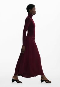Bordeaux ribbet turtleneck top parret med en plisseret maxi nederdel. Modellen bærer sorte hæle sandaler, og fremhæver en elegant silhuet.
