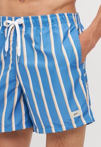 Blue swim shorts made of lightweight fabric featuring vertical stripes in white and orange, elastic waistband with white drawstring and side pocket.