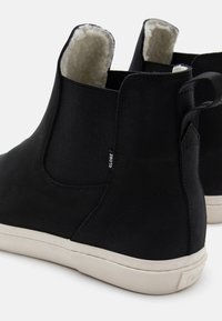 Botines de ante negros con paneles laterales elásticos, forro de piel de oveja color crema y suela de goma blanca. Presenta una pequeña etiqueta de logo en el talón.