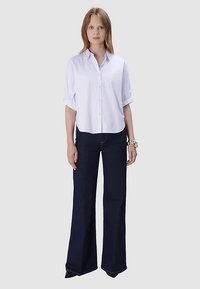 Chemise blanche boutonnée avec manches retroussées, associée à un jean évasé bleu foncé. Le mannequin se tient devant un fond gris neutre, portant des chaussures noires.