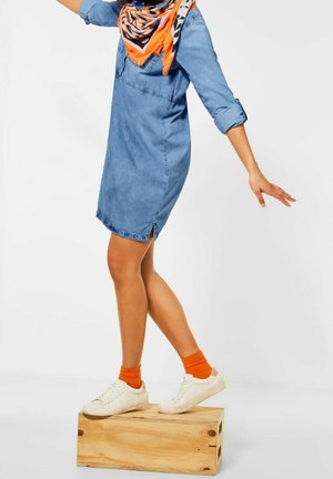 Lichte blauwe denim jurk met opgerolde mouwen en zij-splitten. Gecombineerd met witte sneakers en feloranje sokken, staande op een houten blok.