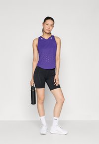 Haut de sport sans manches violet avec design perforé, associé à un short de cyclisme noir. Gourd deau noir ; des chaussettes blanches et des baskets complètent le look.