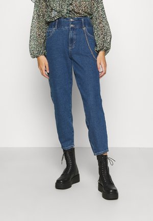 Persoon draagt high-waisted blauwe jeans met kettingdetail, zwarte veterschoenen en een groene bloemen lange mouwen blouse.