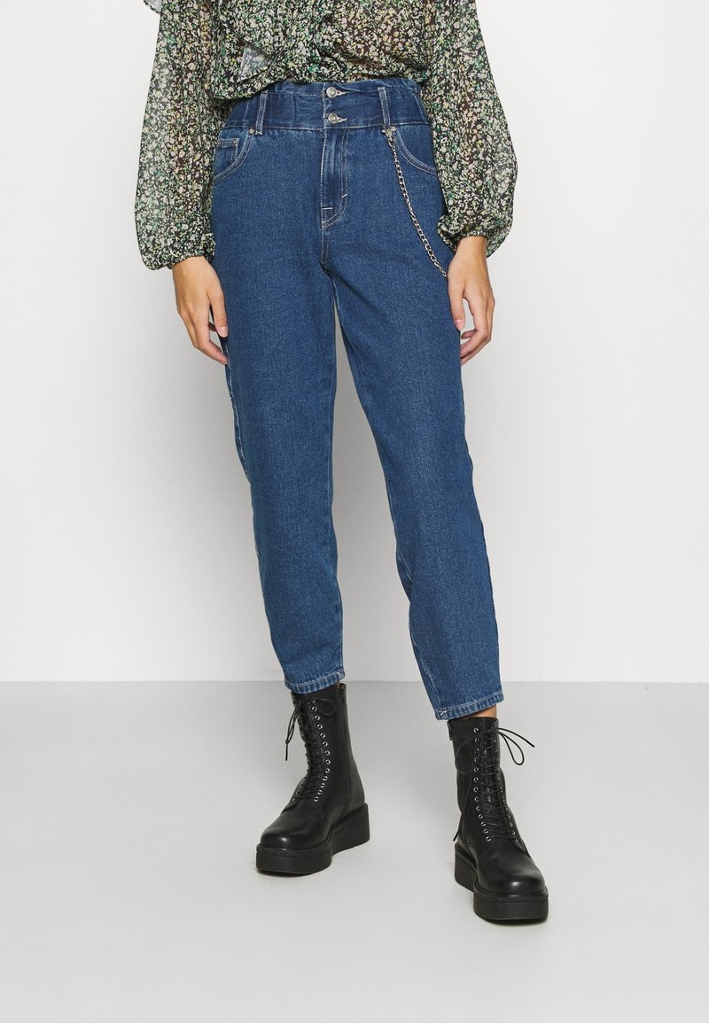 Personne portant un jean bleu taille haute avec un détail chaîne, des bottes noires à lacets et un chemisier vert à motifs floraux à manches longues.