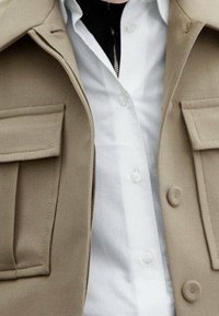 Veste d'extérieur beige avec deux grandes poches à rabat, fermeture à boutons et une chemise blanche bien repassée en dessous. Détails de tissu texturé visibles.
