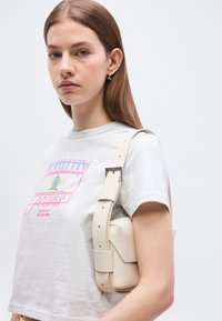 Νεαρή γυναίκα με μακριά καστανά μαλλιά, φορώντας ανοιχτό γκρι μπλουζάκι Tommy Jeans και κρατώντας μια μικρή μπεζ τσάντα ώμου.