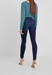 Jeans ajustados de mezclilla azul oscuro, con una cintura de altura media, corte slim, bolsillos traseros y un sutil detalle de costura en la parte posterior. Combinados con una blusa de color teal y tacones beige.