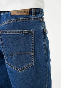 Vaqueros de mezclilla azul oscuro con costuras naranjas, que presentan una etiqueta de cuero en la cinturilla y una etiqueta en el bolsillo trasero. Diseño clásico.