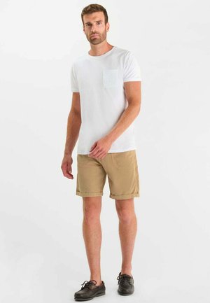 Uomo che indossa una maglietta bianca a maniche corte con taschino, pantaloncini beige fino al ginocchio e scarpe stringate marrone scuro, in piedi contro uno sfondo semplice.