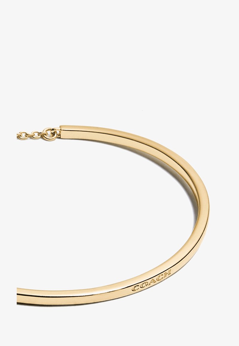 Gold bangle bracelet with a smooth, rounded shape and polished finish. Features a subtle engraved logo on the edge. Adjustable clasp.