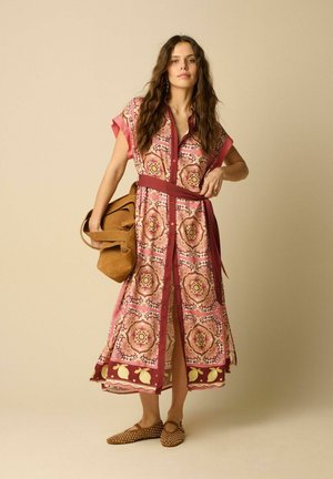 Femme aux longs cheveux porte une robe ceinturée rose et rouge à motifs, des ballerines marron cloutées et tient un grand sac en daim beige.