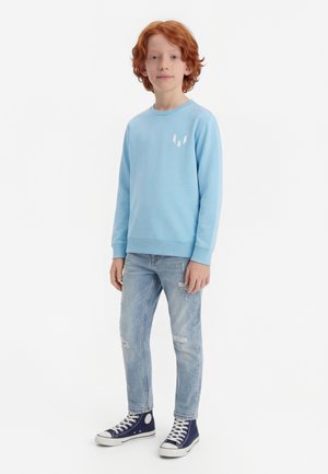 Κόκκινοξανθος αγόρι που στεκεται φορώντας ένα ανοιχτό μπλε φούτερ, σκισμένα ανοιχτόχρωμα τζιν και ναυτικά μπλε ψηλά αθλητικά παπούτσια σε λευκό φόντο.