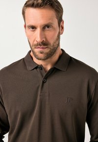 Camisa polo marrón en una tela texturizada con cuello, tres botones y un logo bordado en el lado izquierdo del pecho.