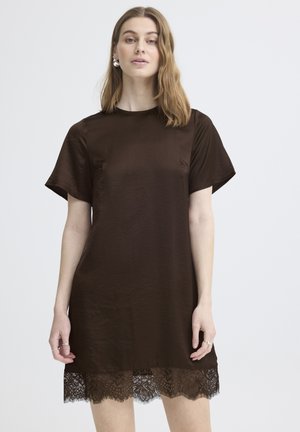 Femme portant une robe marron foncé à manches courtes avec un bord en dentelle à l'ourlet, debout devant un fond clair uni.