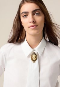 Chemise blanche à col avec une cravate, ornée d'une broche en ton doré représentant un profil sculpté dans un cadre orné, décoré de petites touches sombres.