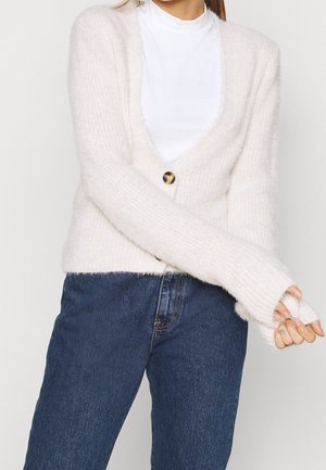 Frau trägt eine weiße, flauschige Strickjacke mit einem Knopf über einem weißen Hemd und blauer Jeans, die Hände vor dem Körper verschränkt.