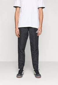 Άτομο που φοράει μαύρο παντελόνι φόρμας Adidas, λευκό μπλουζάκι και ασπρόμαυρα αθλητικά παπούτσια, στέκεται μπροστά σε ένα απλό φωτεινό φόντο.