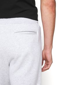 Ljusgrå sweatpants med en slät yta, som har en enda bakficka och ett åtsittande midjeband. En hand vilar nära fickan.