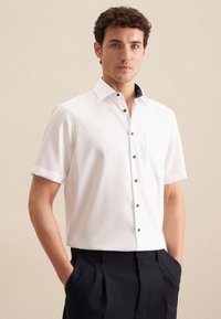 Korte mouwen witte blouse met een zwarte afgewerkte kraag en zak, gemaakt van gladde stof met donkere knopen.