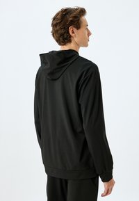 Sudadera con capucha negra de textura suave; presenta mangas largas y un ajuste relajado; diseño minimalista sin patrones visibles.