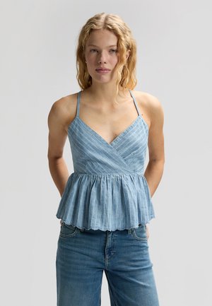 Ung kvinne med krøllete blondt hår iført blåstripet ermeløs peplumtopp og blå jeans, står foran en lys grå bakgrunn.