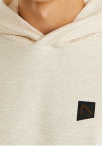 Sweat à capuche beige clair texturé avec une capuche douillette, présentant un petit patch noir avec un logo orange sur la poitrine.