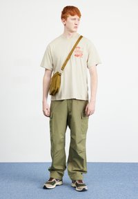 Beige cotton t-shirt with red design, olive green cargo pants, and multi-colored sneakers. Brown sling bag worn crossbody.