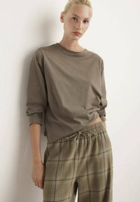 Bruine lange mouwtop gecombineerd met geruite, beige en groene broek met een elastische taille en trekkoord. De stof lijkt zacht en ontspannen.
