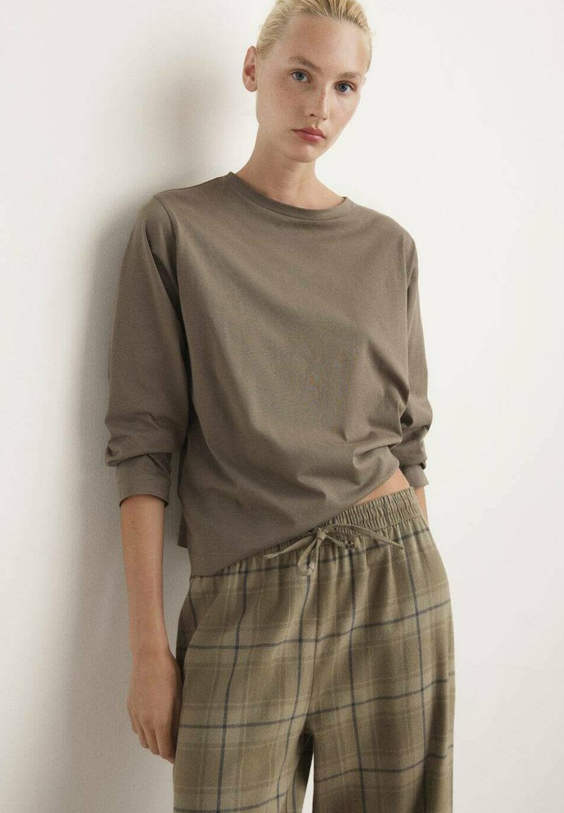 Bruine lange mouwtop gecombineerd met geruite, beige en groene broek met een elastische taille en trekkoord. De stof lijkt zacht en ontspannen.