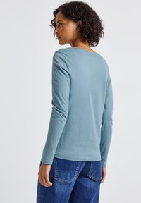 Žena s kudrnatými tmavými vlasy, oblečená v světle modré košili s dlouhým rukávem a modrých džínách, otočená mírně doleva před jednobarevným pozadím.