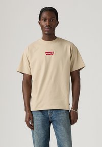 Beige bomull t-skjorte med korte ermer og rund hals, med et rødt logo-patch på brystet og en avslappet passform. Bæres med blå jeans.