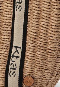 Bolsa de paja tejida con un acabado natural texturizado, presenta una correa negra con letras blancas y una pequeña etiqueta de cuero estampada con "Kbas".
