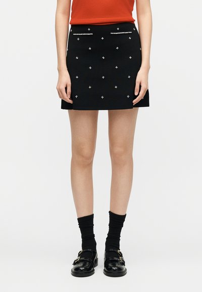 Γυναίκα που φοράει μαύρη μίνι φούστα με μικρές διακοσμητικές καρφίτσες, μαύρες κάλτσες, μαύρα παπούτσια με αγκράφα και κόκκινο αμάνικο τοπ, στέκεται μπροστά σε λευκό φόντο.