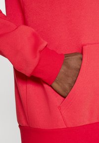 Κόκκινο φούτερ με τσέπη, με απαλή υφή και μανσέτες με ριμπ ύφανση. Στο κοντινό πλάνο φαίνεται ένα χέρι που ακουμπάει μέσα στην τσέπη.